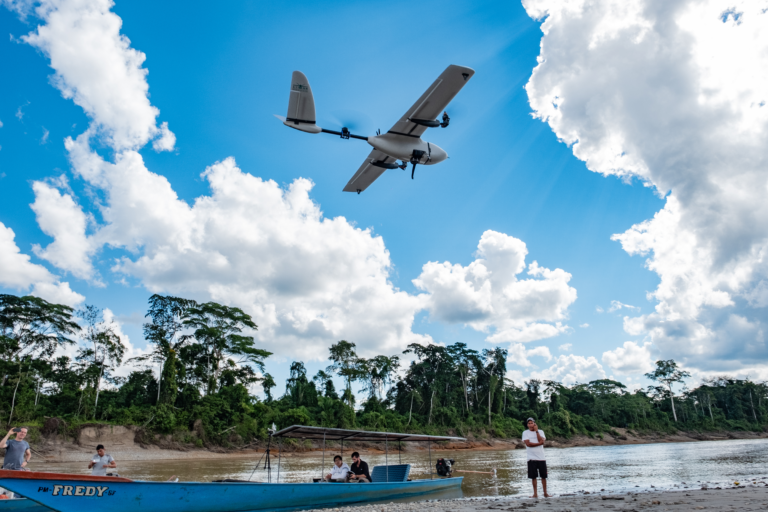 Image of a group of people on a boat in a river basin lined by tall trees. A large drone controlled by one of the group flies overhead.