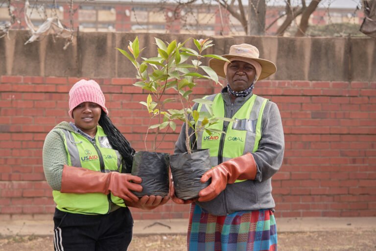 Two women stand proudly with two berry trees as they engage in a reforestation project