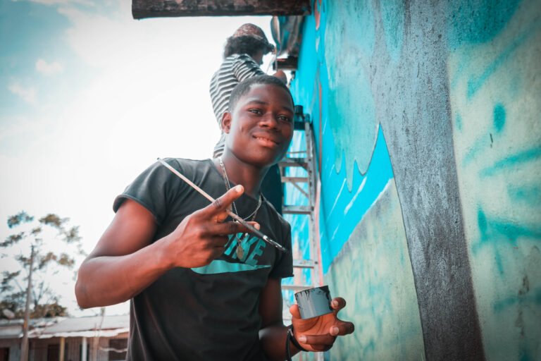 Youth holding paintbrush with hand in a peace sign standing next to wall covered in a painted image