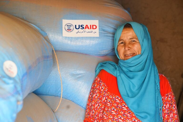 A Moroccan woman wearing a blue hijab poses, smiling at the camera, before sacks of grain that bear the USAID identity.