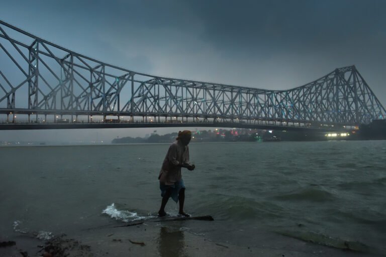 A rain-soaked scene along the banks of the Ganges in Kolkata. The river flows with a vibrant green hue under dark clouds, while the iconic Howrah Bridge towers in the background. A lone figure stands in the rain, embodying the city's resilience.