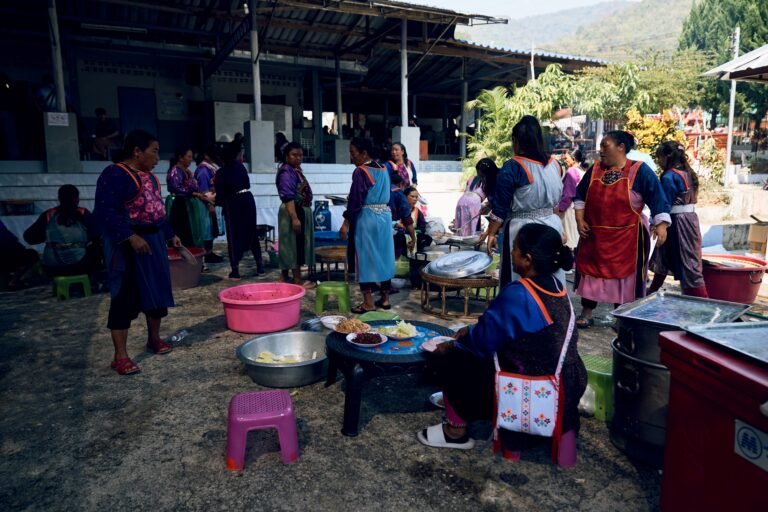 A group of women from the Lisu ethnic group are gathered around cooking utensils and tables. They are and preparing food while wearing traditional clothes.