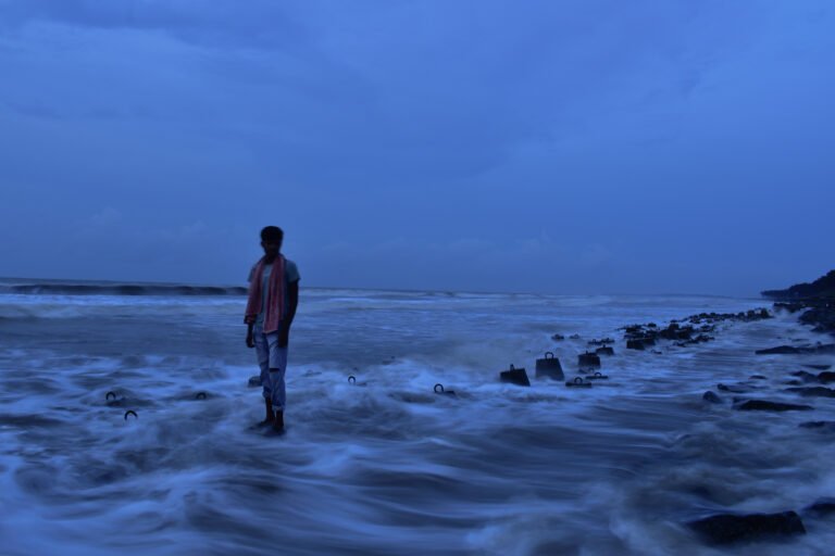 : In this photograph captured along the sea-side of India, a resilient man stands on a boulder as the rising tide gently approaches, depicting the impact of climate change on coastal communities. The blueish tone of the image adds to the serene ambiance, while highlighting the significance of rising sea levels and the challenges faced by the coastal residents. The photograph is part of the series "The Rising Tide," documenting the human spirit's battle against the increasing threats posed by climate change