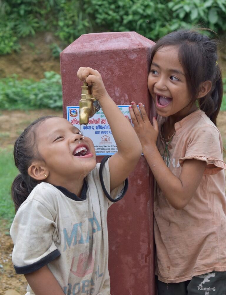 Two children smile as they drink from a water spigot.