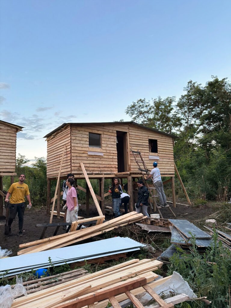 A group of volunteers work together to build a house on stilt with planks of wood in the forefront of the image.