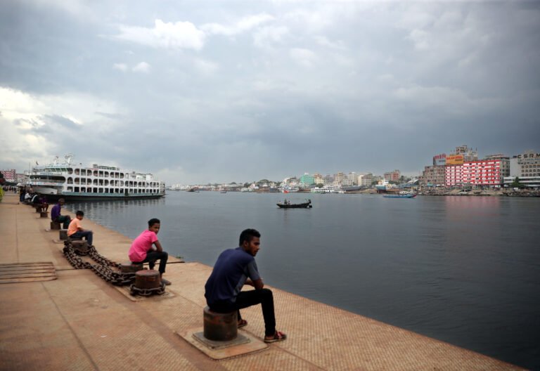 People sit on concrete looking out over a body of water in an urban setting as storm clouds darken the sky overhead.