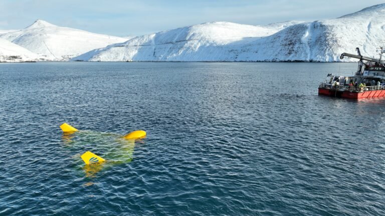 A yellow machine that resembles a kite is partially submerged in a body of water with snow-capped mountains in the background.