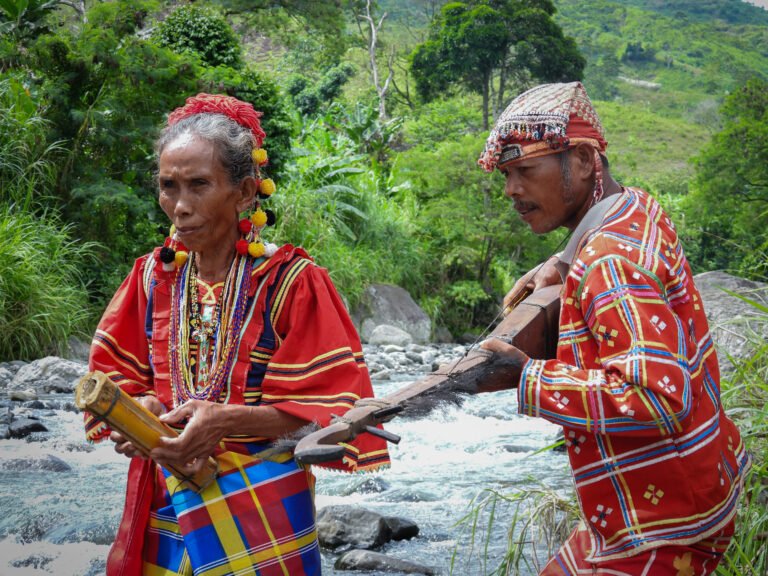 Two elders in traditional red outfits play musical instruments in front of a roaring river surrounded by forest.