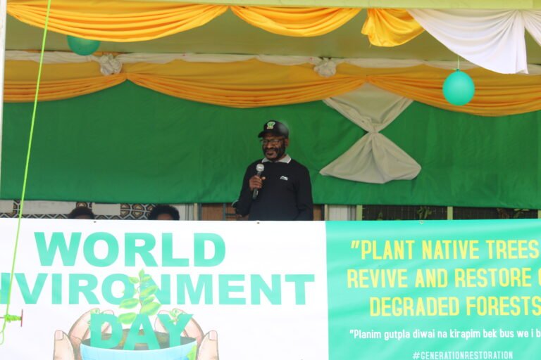 A man holds a microphone while standing on a decorated state with a World Environment Day banner in front of him.