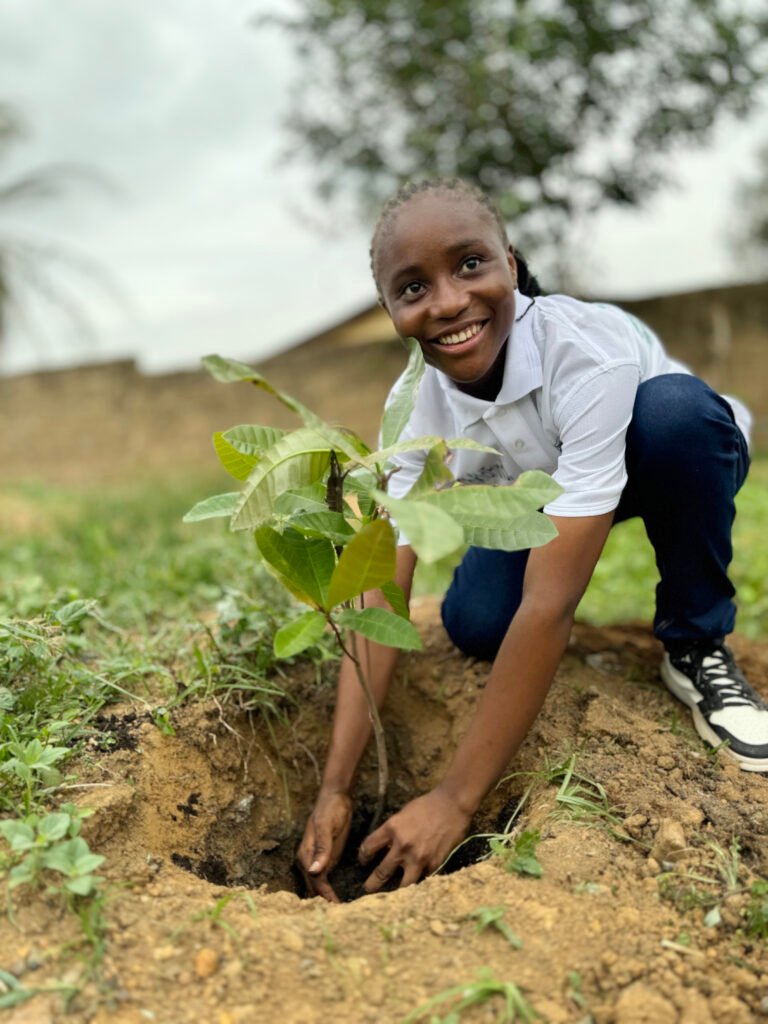 A child crouches down to plant a tree sapling while smiling at the camera.