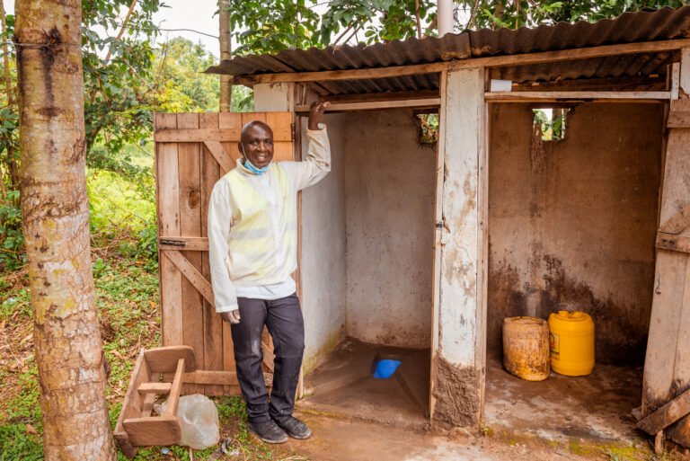 A man stands smiling in front of a latrine.