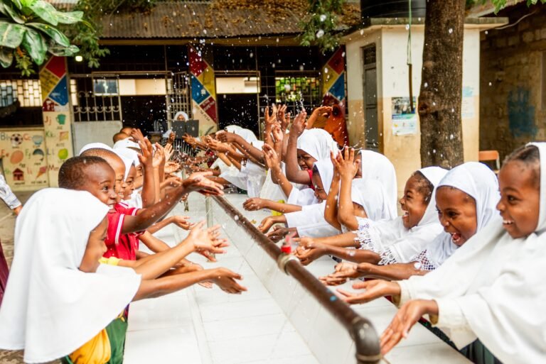 A group of more than 15 elementary school-aged students line up at a long sink facing each other as they laugh and splash water at each other from a long pipe.