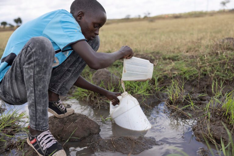 A young boy crouching down to collect water from a muddy stream.