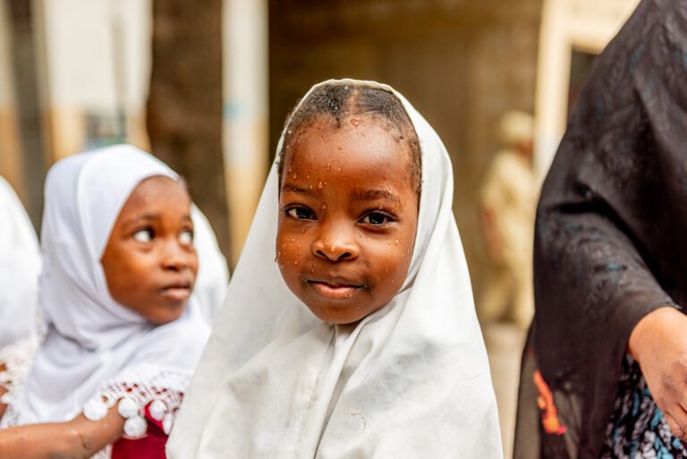 A young girl wearing a white headscarf, with water droplets on her face, smiles at the camera.