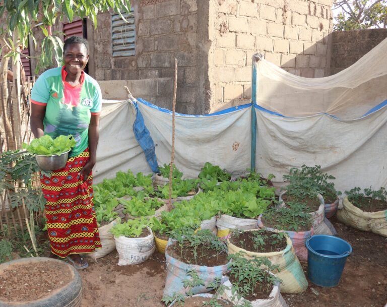 A woman smiles as she shows off her many container plants outside her brick home.