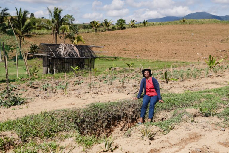 A woman sits on her farm, with a small wooden structure and mountains visible in the distance.