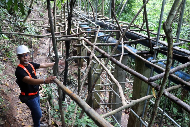 A man in an orange safety hat and white hard hat repairs a large piping system in the forest.