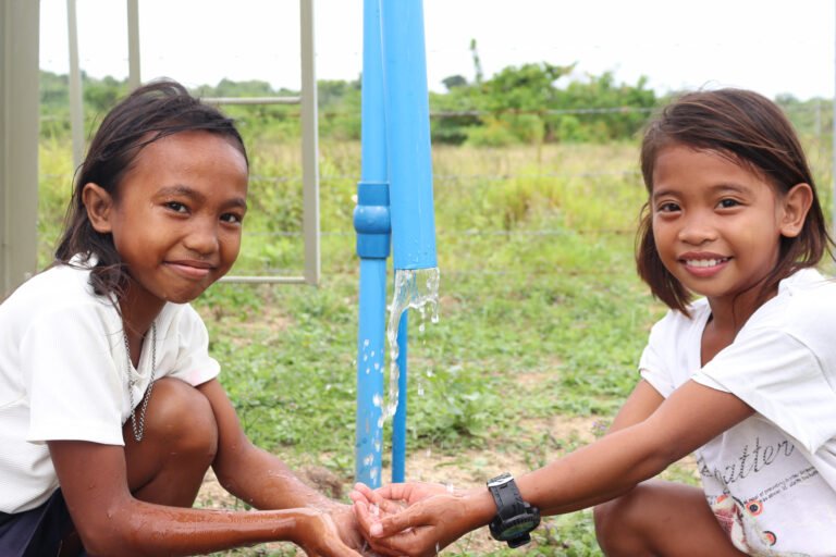 Two young girls smile at the camera as they cup their hands under a running outdoor tap.