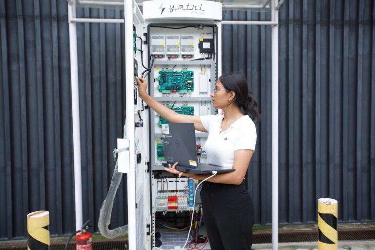 A woman holds a laptop in one hand while using the other to fix wires in a large open electric vehicle charging station.