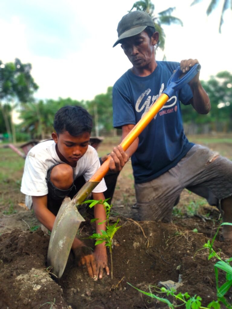An adolescent boy bends to plant a tree sapling as an older man helps with a shovel.