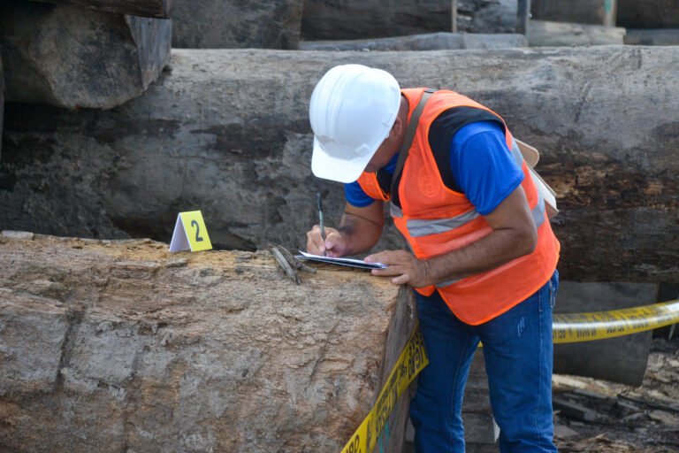 A man in an orange safety vest and white hard hat leans on a fallen log to take notes on paper.