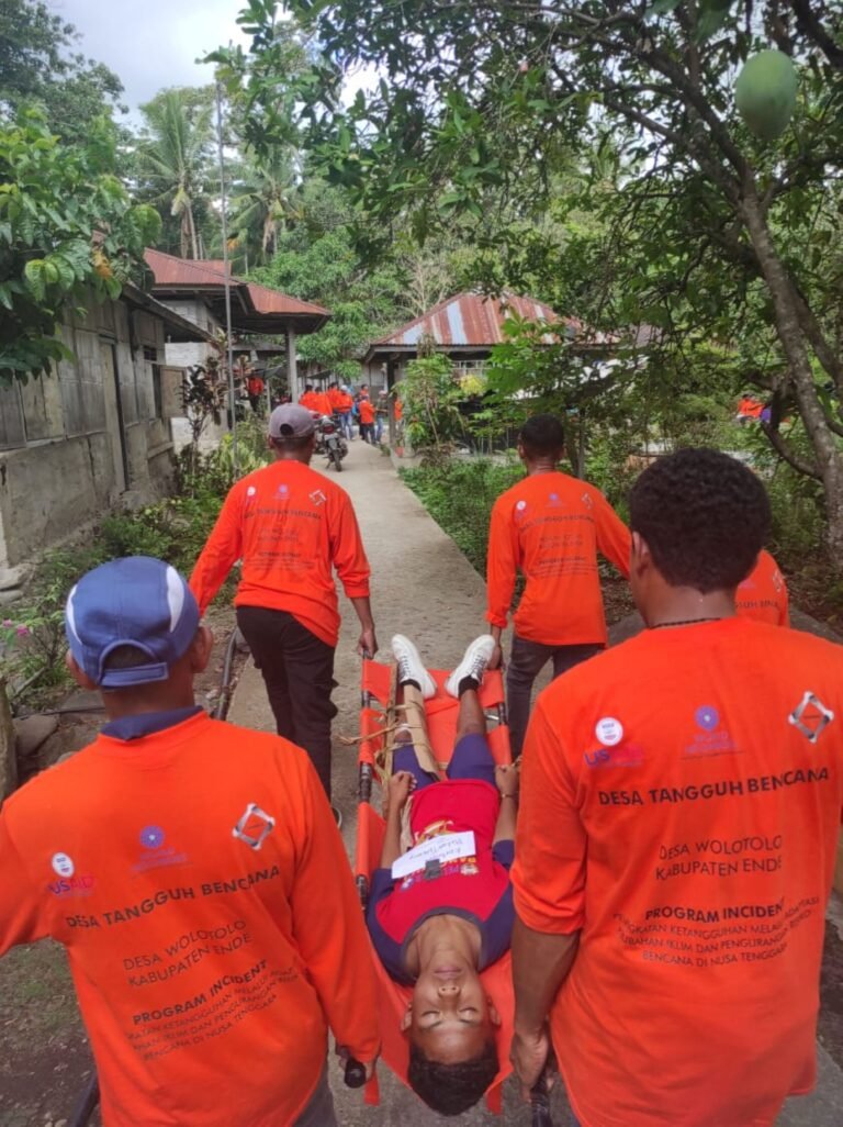 People in red carry a person lying on a stretcher through a village street.
