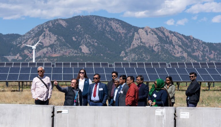 Partners from Bangladesh learning about solar and wind turbines outdoors with solar panels, a wind turbine, and mountains in the background..