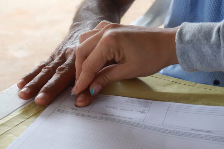A person's hand presses another’s index finger to help leave their fingerprint in ink on a piece of paper.