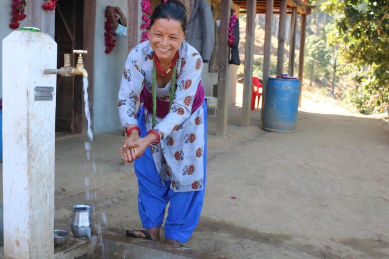 A woman smiling while washing her hands at an outdoor water tap.