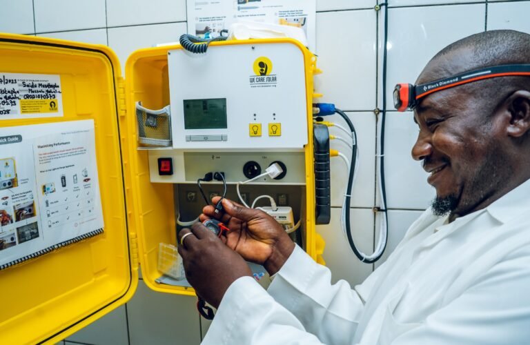 A healthcare worker wearing a white medical coat smiles as he wears a head lamp and fixes wires protruding from a solar suitcase mounted to the wall.