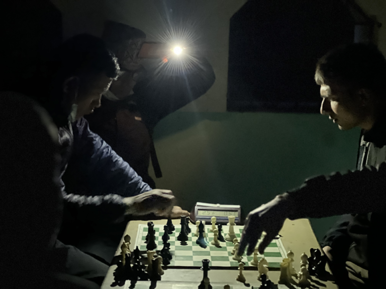 Two chess players and another non player that lights the space with their mobile phone's torch light