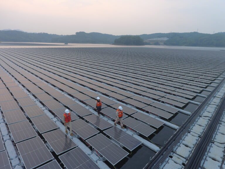 Three people in hard hats walk on a massive floating solar plant, with the rows of solar panels stretching to the body of water and hills behind them.