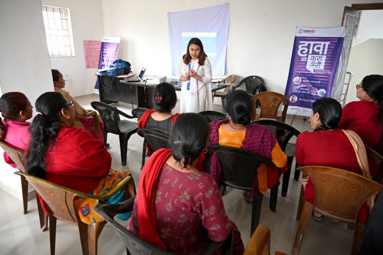 A group of women listen as a woman stands in front of them speaking in a classroom setting.