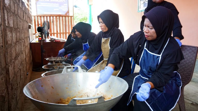 Women in aprons and gloves work side by side over large woks.