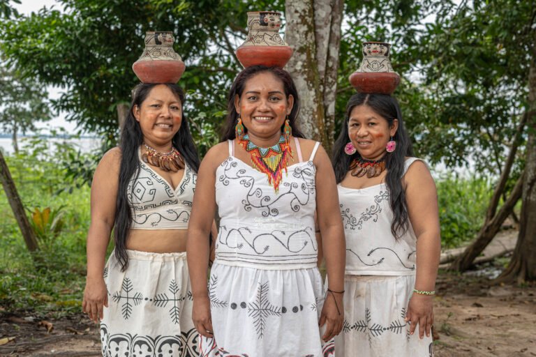 Three Indigenous women wearing beaded jewelry and embroidered outfits smile at the camera as they display pottery on their heads in a forest clearing.