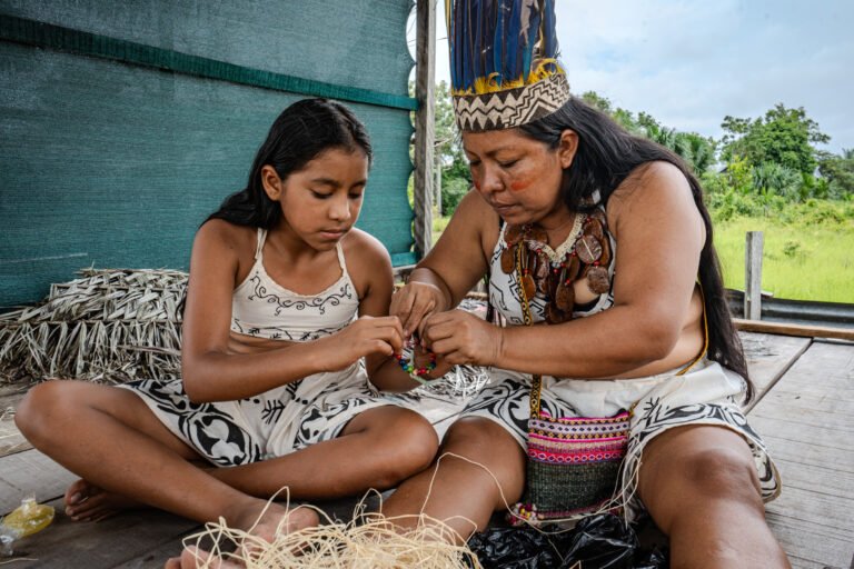 Two Indigenous women sit together on the floor working on a beaded handicraft.