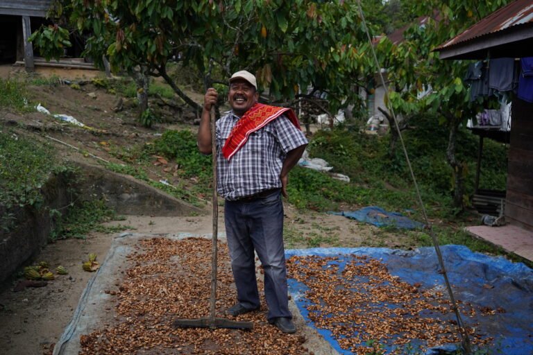 A farmer smiles with a rake as he stands amid coffee cherries outside.