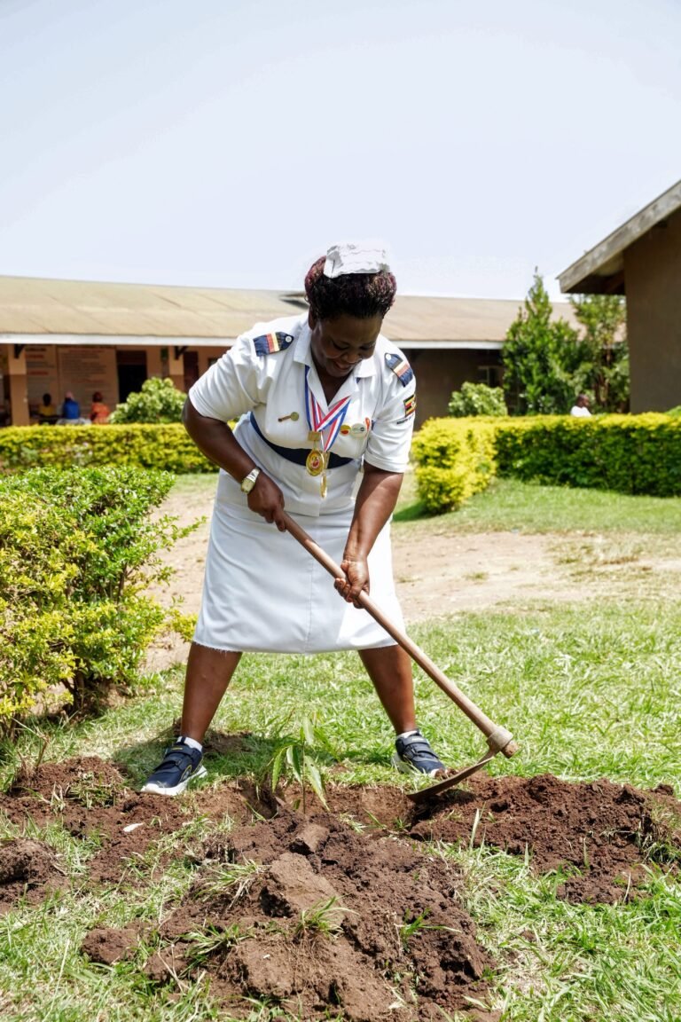 A midwife in a white uniform uses a shovel to move dirt to plant a tree outside her facility.