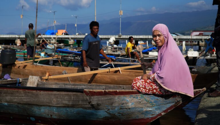 A woman looks at the camera from her small boat as she sits in a busy port.