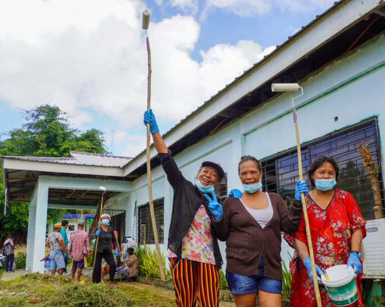 Three women smile as they pose with paint buckets and rollers outside of a building, with other people standing or sitting behind them.