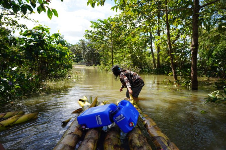 A woman wades through shallow water, dragging two blue tanks on four logs tied together.