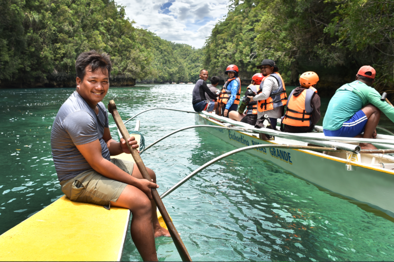 A tour guide on a boat smiles at the camera as he steers a boat full of tourists wearing helmets.