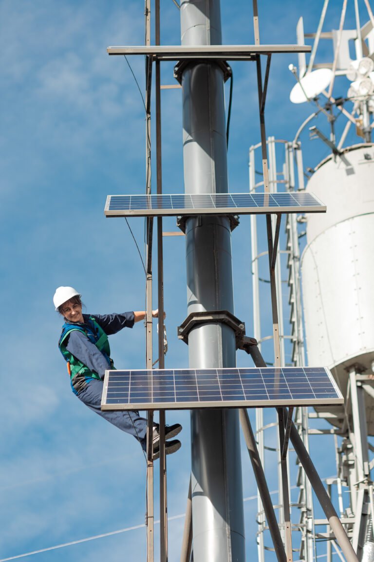A woman in a hardhat smiles as she leans off a ladder on a tall tower outfitted with solar panels.