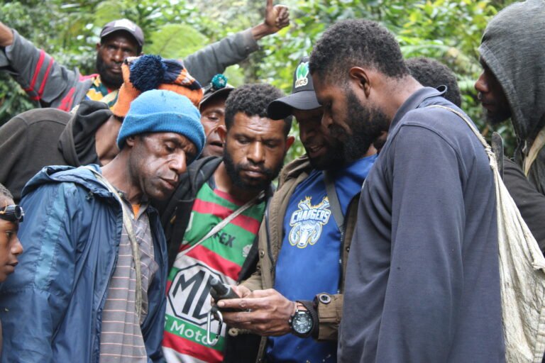 A group of men in the forest huddle together to look at a GPS tool.