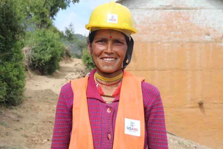 A woman in an orange safety vest and yellow safety helmet smiles at the camera in front of a water source.
