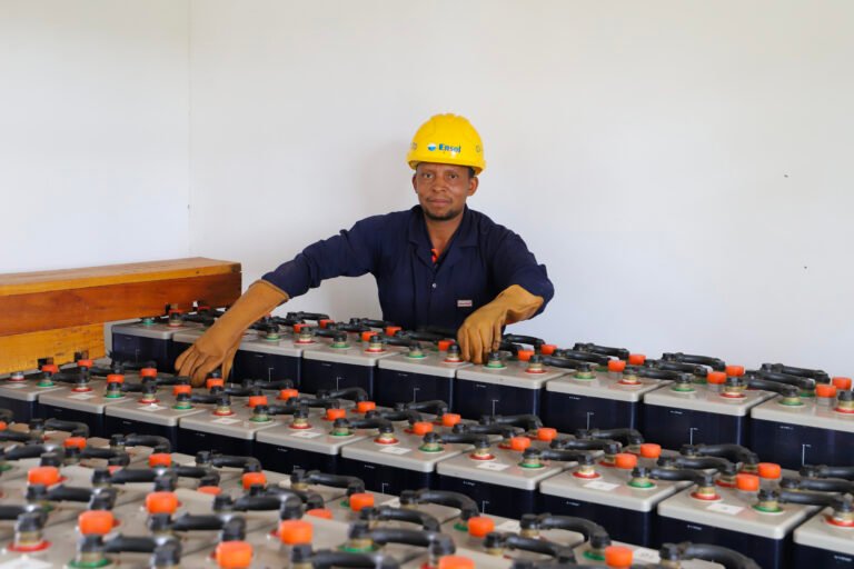A person in a yellow hard hat pauses while working with rows of large batteries.