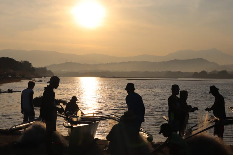 A group of fisherfolk are backlit by the rising sun as they prepare their nets, mountains visible in the hazy background.