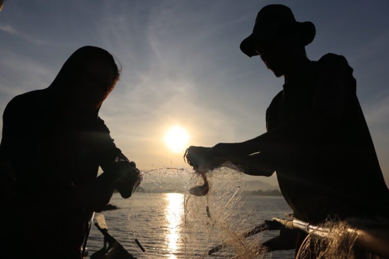 Two fishermen hold up a net between them to count the few small fish in it as the sun illuminates them in shadow.