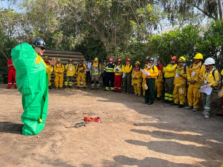 A group of firefighters stand in a semi circle watching an instructor demonstrate how to use a fire shelter.