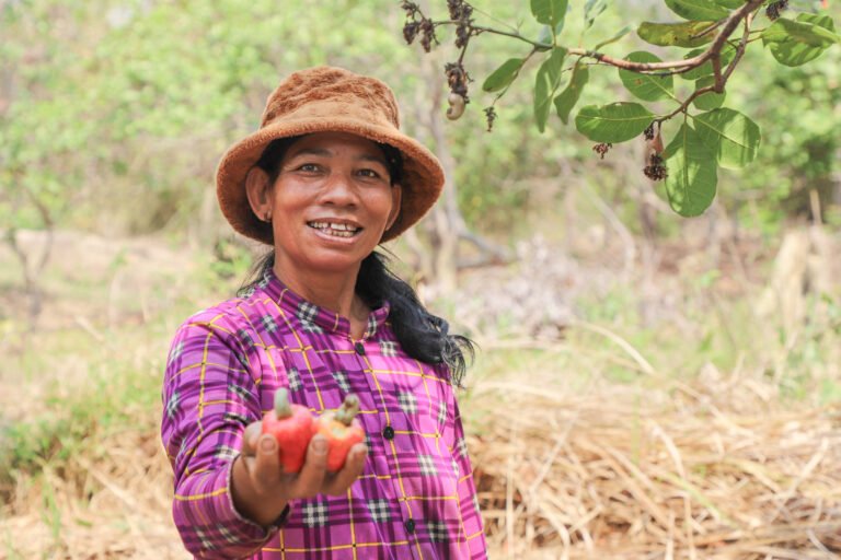 A woman farmer holds two cashew apples in her hand while smiling at the camera.